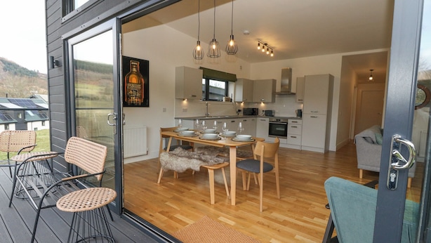 Une cuisine moderne avec des meubles en bois, un coin repas avec une table dressée pour quatre personnes, et une vue sur l’extérieur grâce à de grandes portes vitrées.