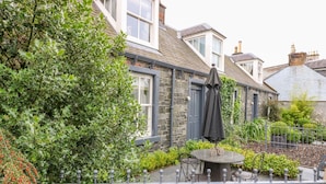 A stone house with a patio area featuring a round table and chairs, surrounded by greenery and a metal fence. - Wee Cottage (Moffat)