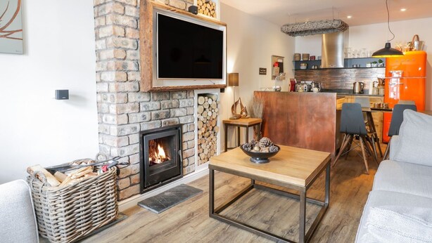A modern living room with a fireplace, a television mounted on a brick wall, a dining area with a table and chairs, and a kitchen area with a refrigerator and counter. - Wee Cottage (Moffat)