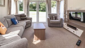 Ein großzügiges Wohnzimmer mit einer Couch, einem Sessel und einem Couchtisch. Ein Fernseher steht auf einem Holzgestell, und eine Glastür öffnet sich zu einem Garten.