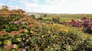 A garden with various plants and flowers, a pathway, and a view of a rural landscape with fields and a distant village. - An Radharc Álainn (Rosscarbery)