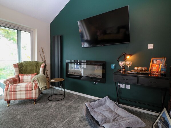 A room with a plaid armchair, a small round table, a flat-screen TV mounted on a green wall, a side table with a lamp, and a pet bed. - The Sail Loft (Y Felinheli)
