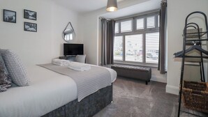 A bedroom with a bed, a TV, a window with blinds, a hanging rack, and a bench. - 119 Henver Road (Newquay)