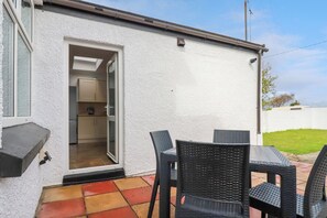 A patio area with a table and chairs, a white exterior wall, and a door leading to a kitchen.