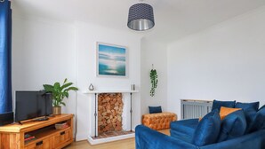 A living room with a blue sofa, a wooden entertainment unit, a fireplace, and a hanging plant. - 119 Henver Road (Newquay)