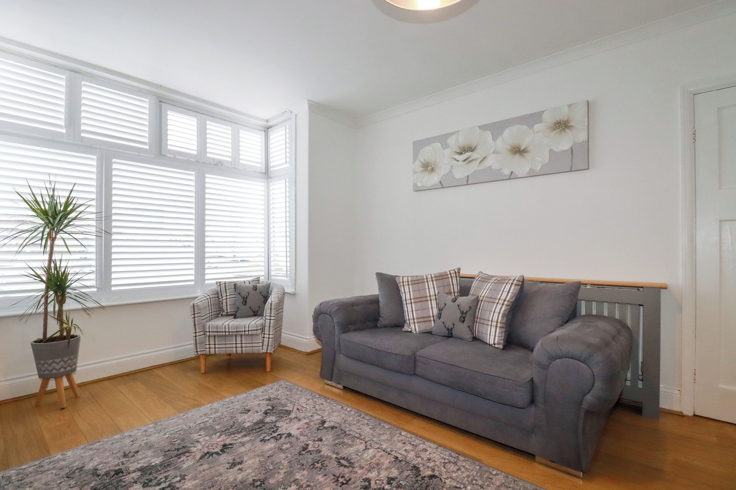 A living room with a grey sofa, a plaid armchair, a large window with blinds, a potted plant, and a floral artwork on the wall.