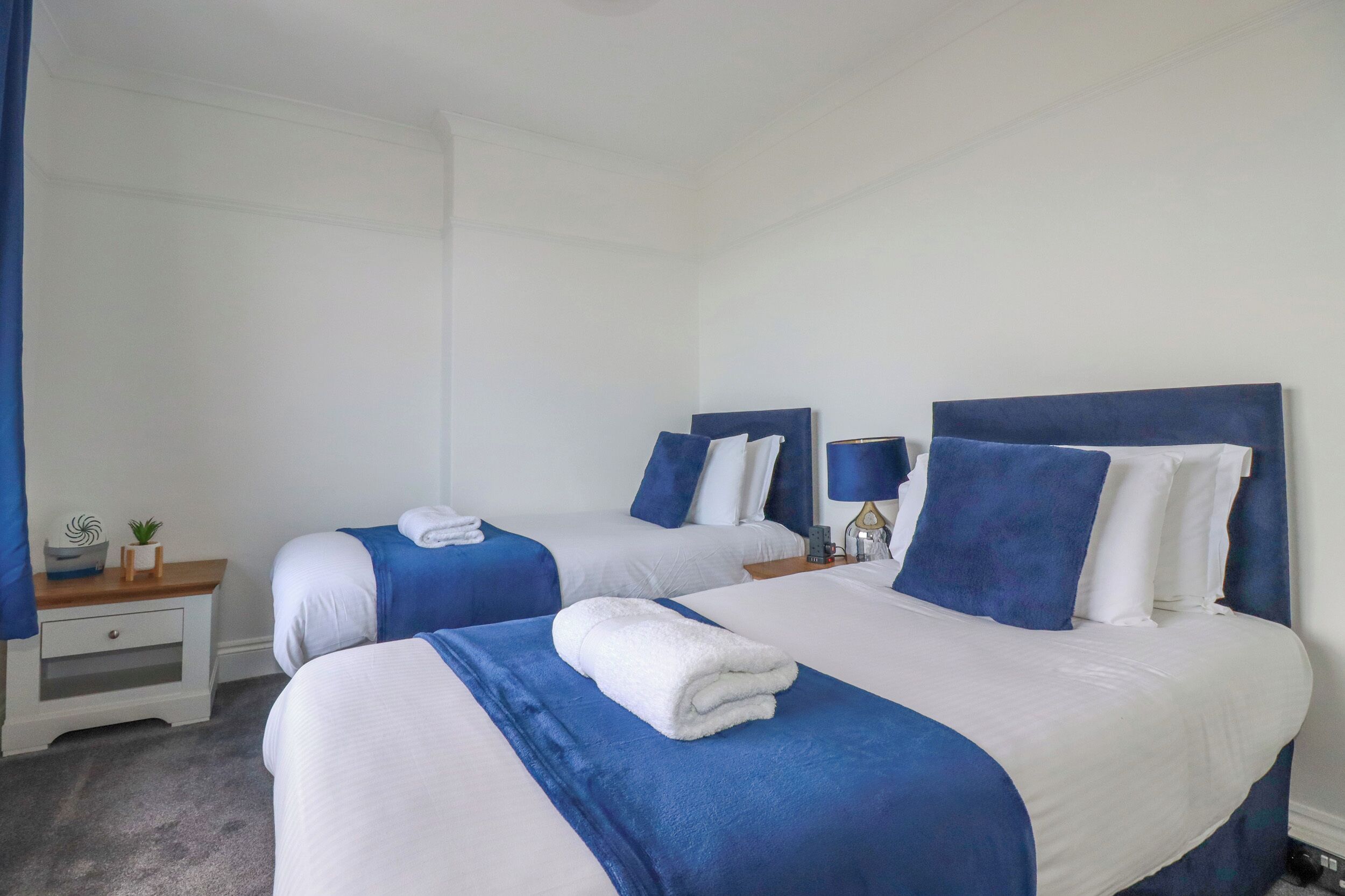 Een hotelkamer met twee bedden, elk voorzien van blauw-witte beddengoed, en een klein nachtkastje met een lamp.