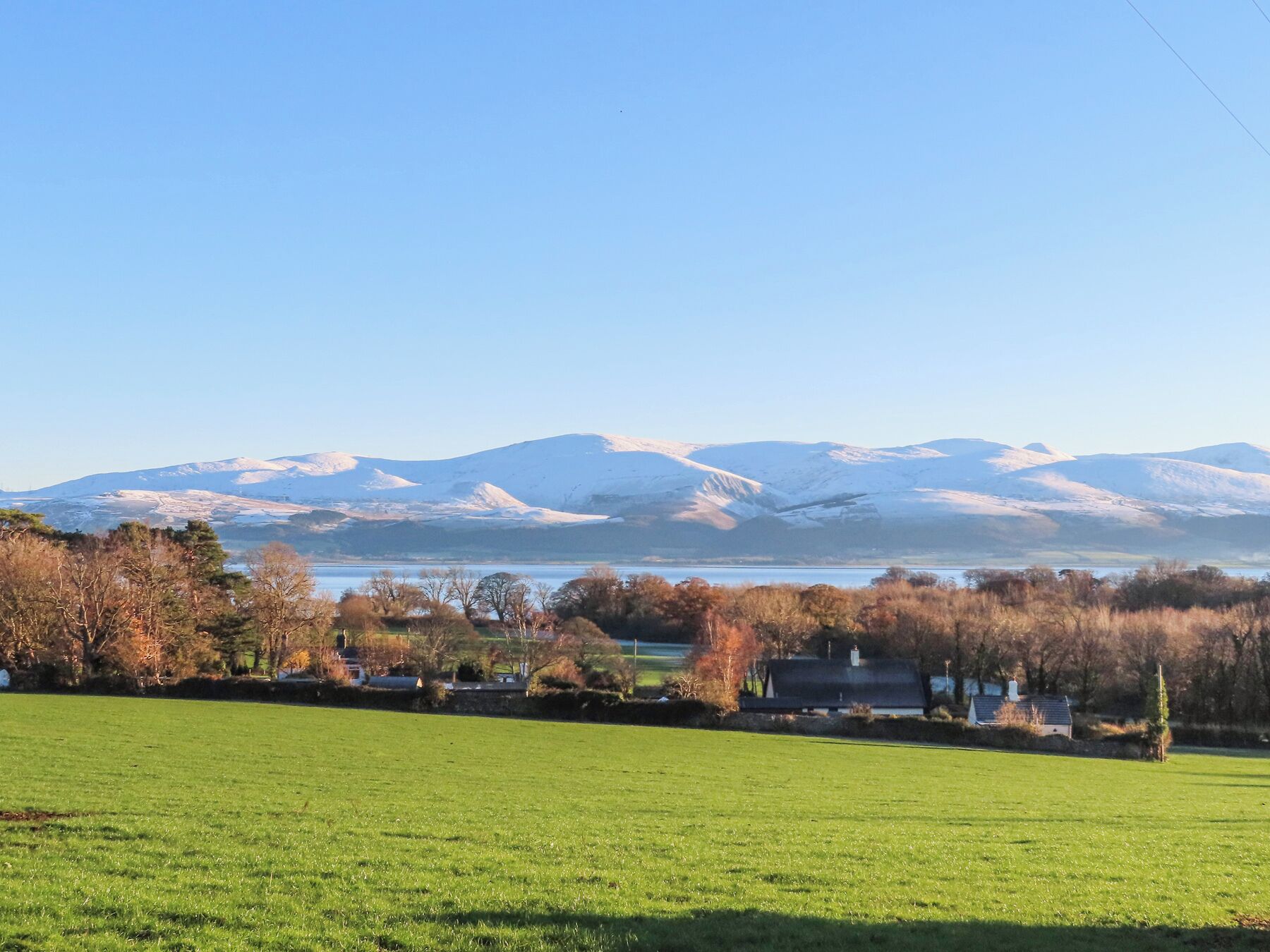 Paisagem rural com um lago, montanhas nevadas e um campo verde.