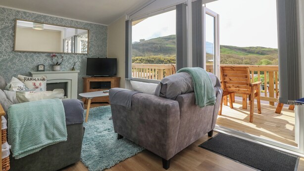 A living room with a sofa, a television, and a fireplace. There is a balcony with a view of the countryside. - Y Goedlan (Swansea)