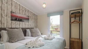 A neatly made bed with a light blue bedspread and white pillows, a wooden headboard, a wall-mounted picture, a hanging light fixture, and a wooden side table with a hanger. - Y Goedlan (Swansea)