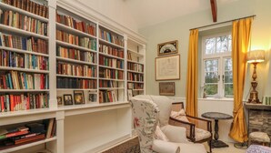 Una biblioteca con estanterías, un sillón de lectura, una pequeña mesa y un ventanal con cortinas.