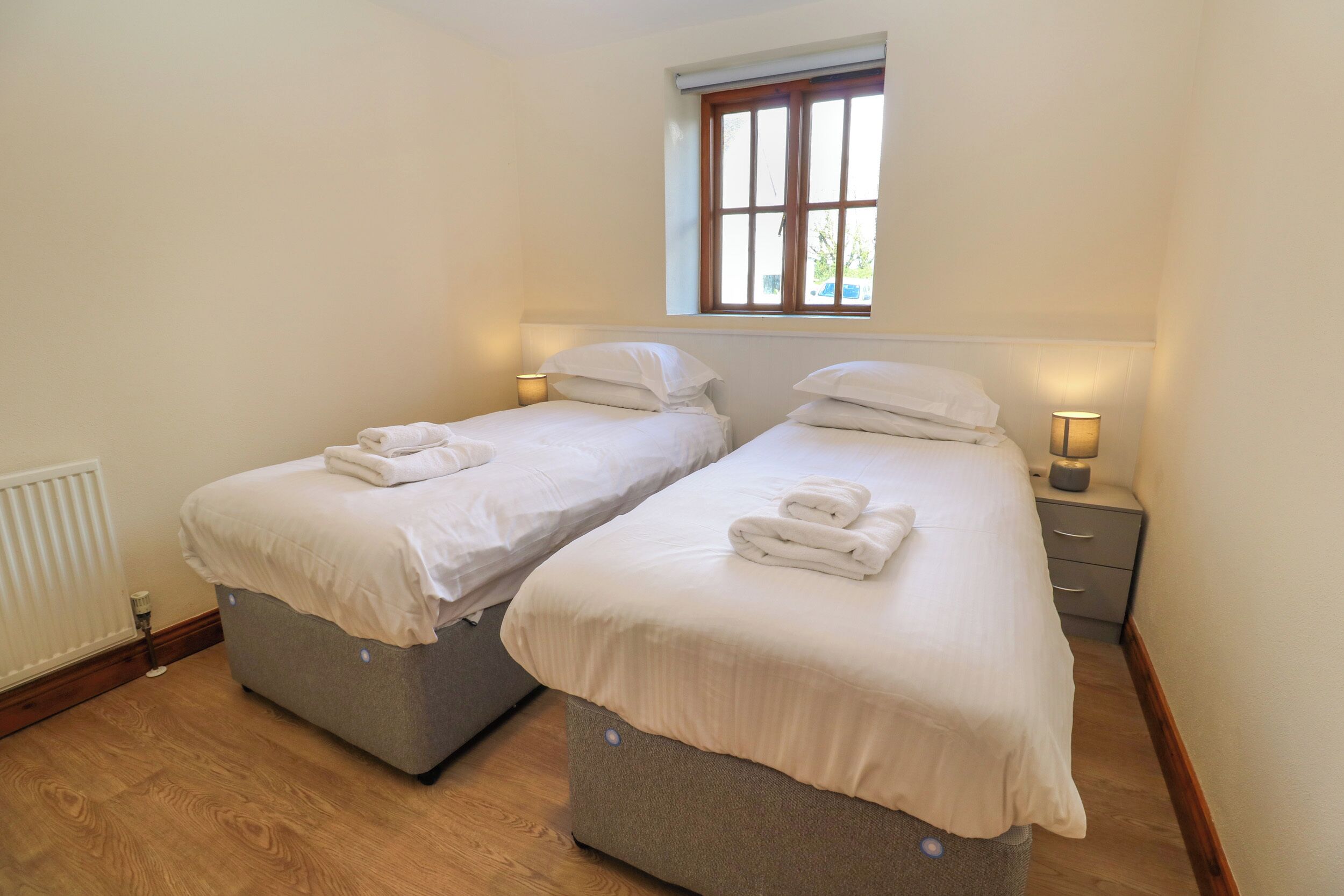 Un dormitorio con dos camas individuales, cada una con una mesita de noche y lámpara, una ventana sobre las camas y un radiador en la pared.