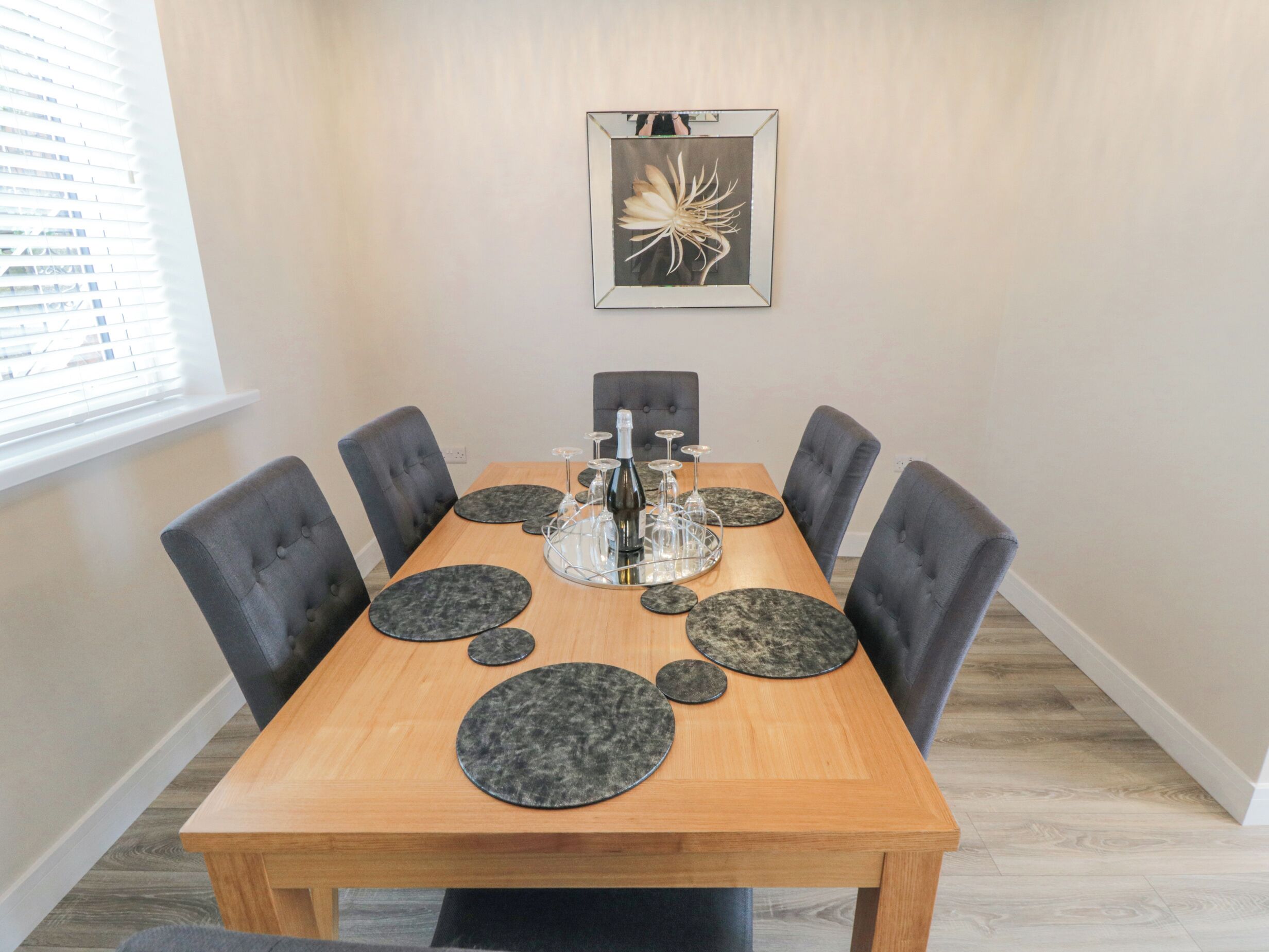 A dining table with six chairs, a centerpiece with a bottle and glasses, and a framed artwork on the wall.
