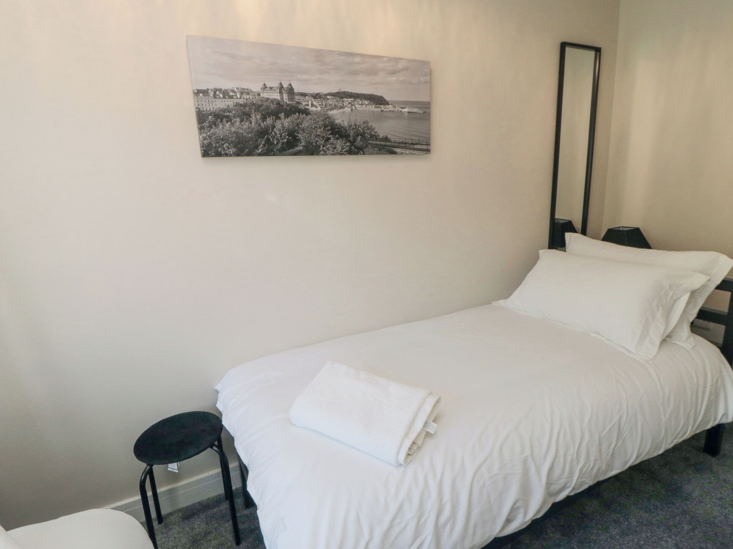 A neatly made bed with white linens, a black chair, and a framed black and white photograph on the wall.