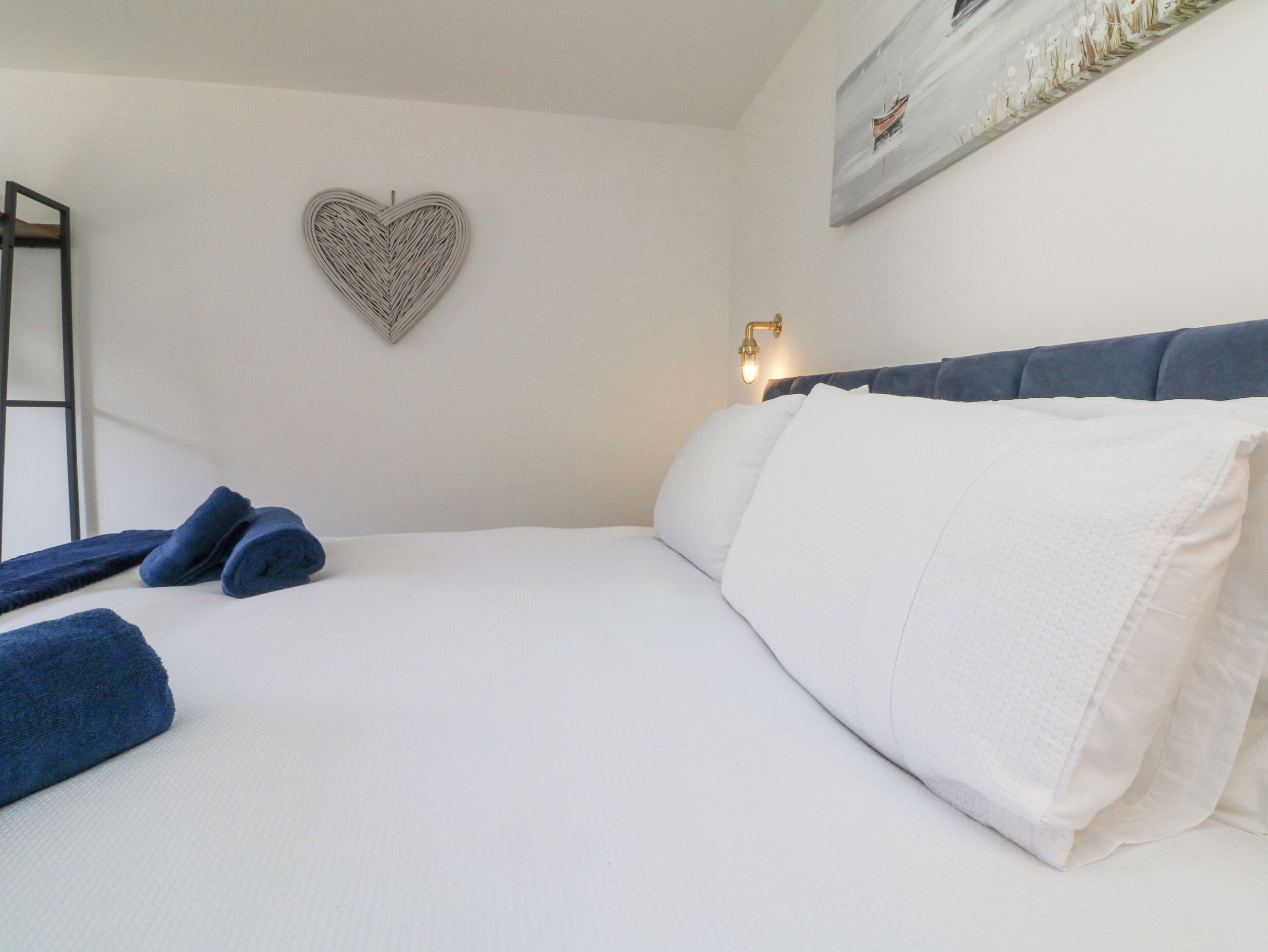 A neatly made bed with white linens and blue towels, a heart-shaped wall decoration, and a painting of a boat on the wall.