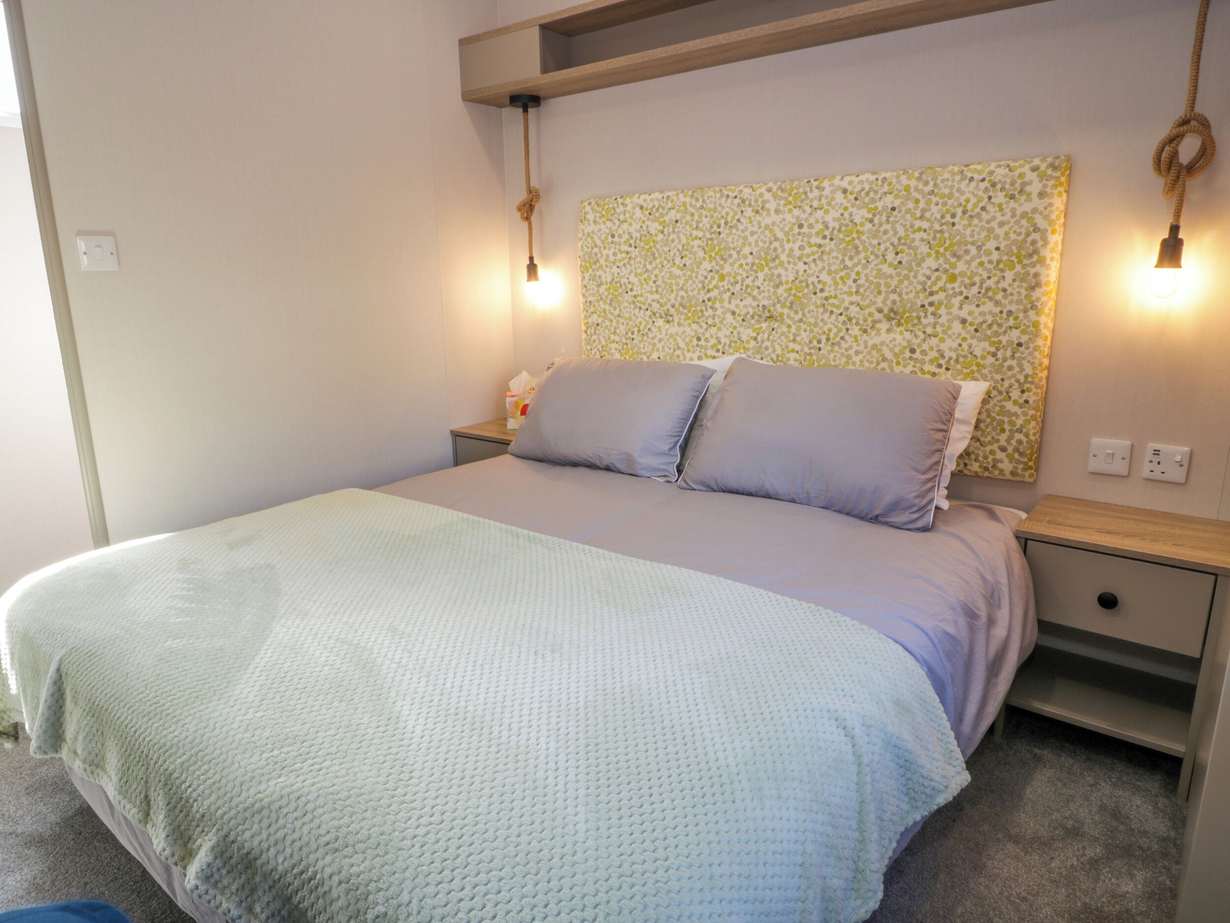 A bedroom with a bed, two bedside tables, a nightstand, a wall-mounted light fixture, and a power outlet.