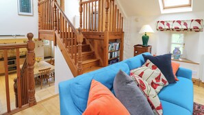 Ein geräumiger Wohnbereich mit einer Holztreppe, einem blauen Sofa mit vielen Kissen, einem Bücherregal und einem Fenster mit einer dekorativen Gardine.