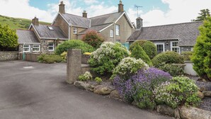 A stone house with a driveway leading to a gated entrance, surrounded by well-maintained gardens with various shrubs and flowering plants. - Ty Llaeth Cottage (Tywyn)