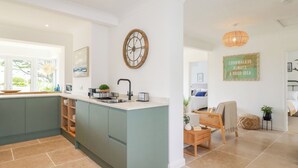 A modern kitchen with a large island, a clock on the wall, and a cozy seating area with a view of the outdoors. - Polwen (Padstow)