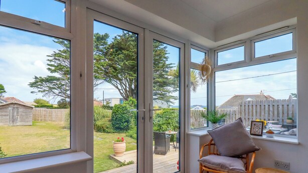 A conservatory with large windows, a wicker chair, a small table, and a view of a garden and neighboring houses. - Polwen (Padstow)