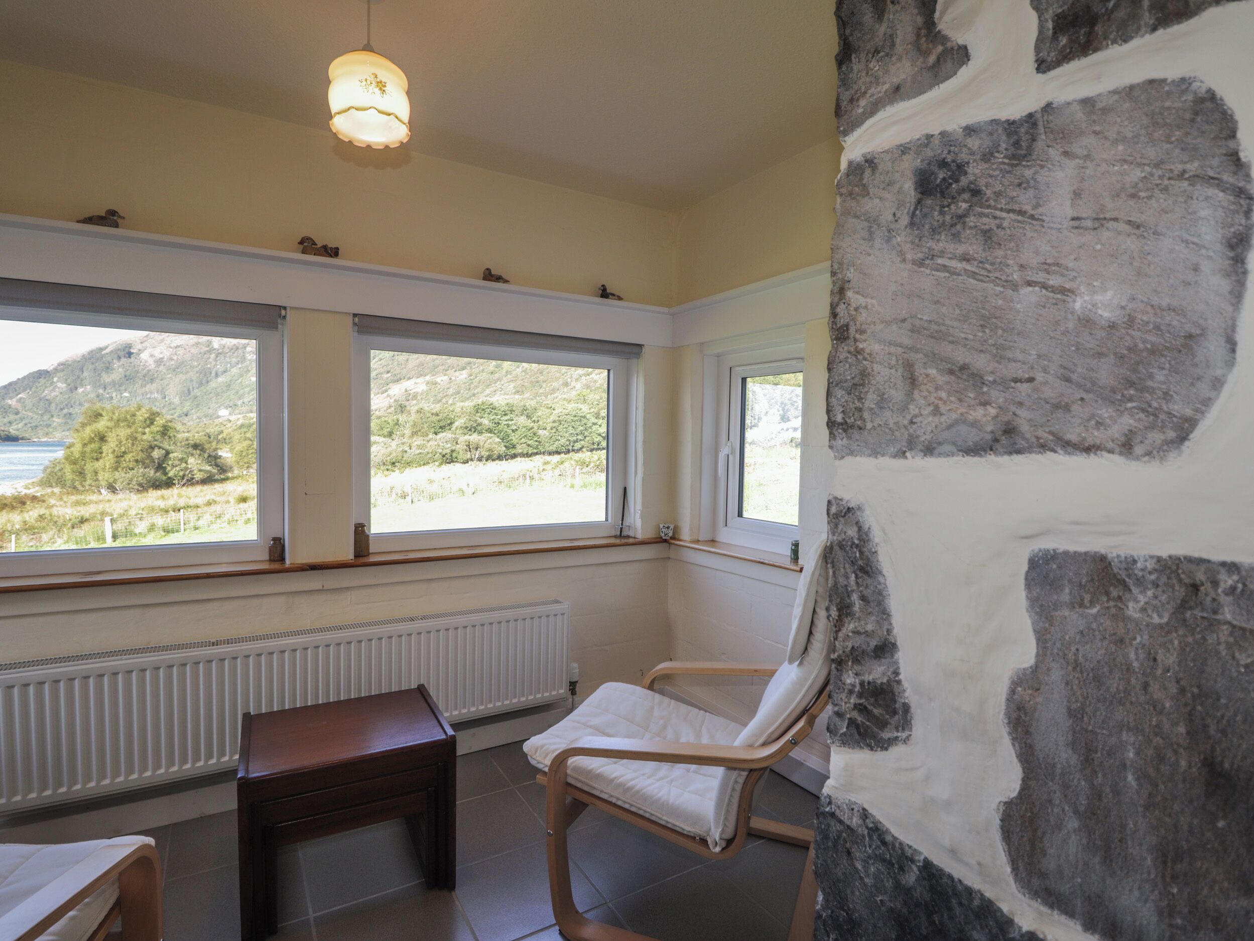 Una habitación con pared de piedra, una mesa de madera, una silla blanca y vistas al paisaje a través de amplios ventanales.