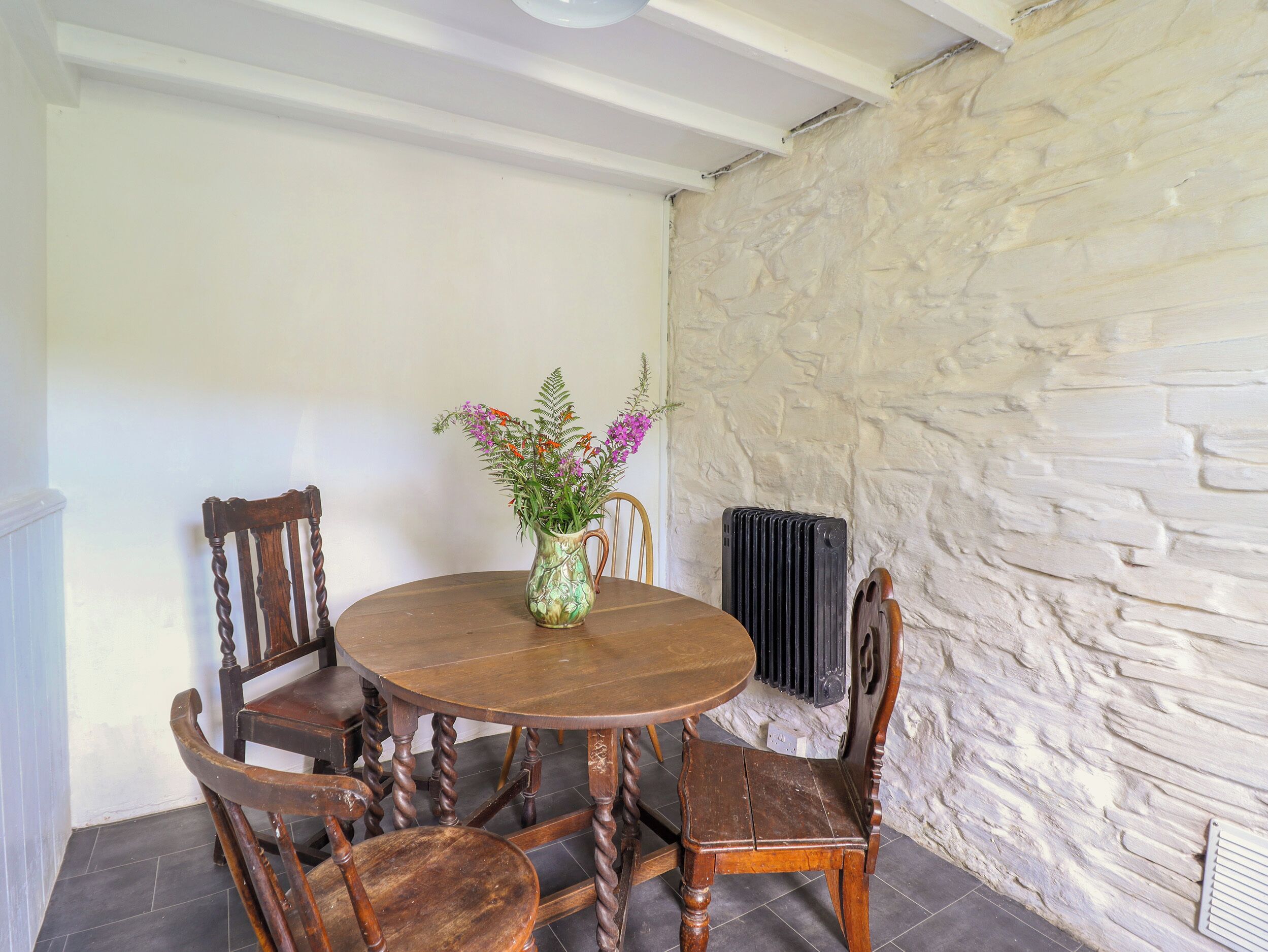 A round wooden dining table with a vase of flowers, surrounded by wooden chairs with carved details. A stone wall and a radiator are visible in the background.