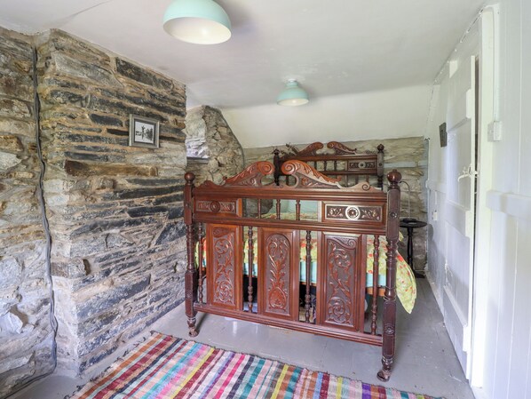 A bedroom with a stone wall, a wooden bed with intricate carvings, a colorful striped rug, and a ceiling light fixture.