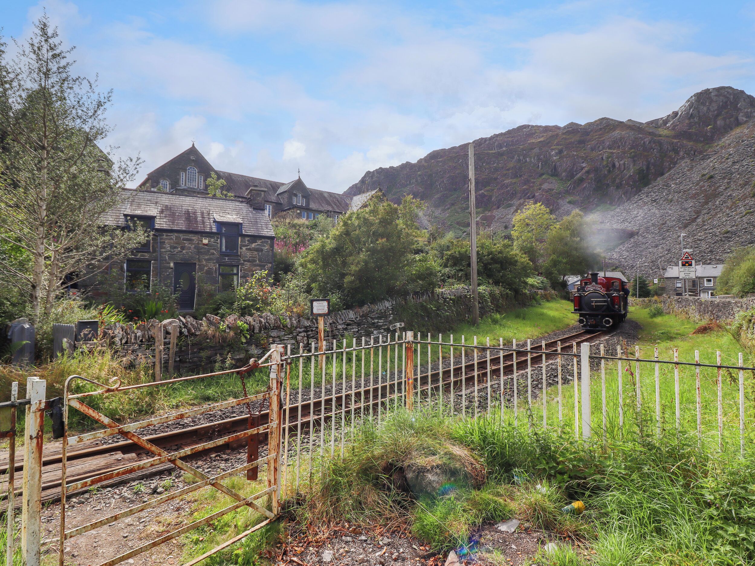 Une voie ferrée pittoresque, empruntée par un train à vapeur, entourée d’une végétation luxuriante et bordée d’un bâtiment en pierre.