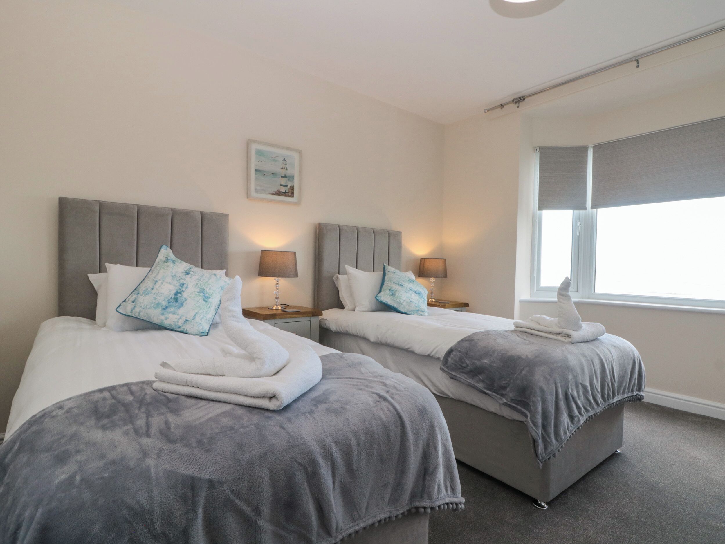Habitación de hotel con dos camas, cada una con cabecero gris, ropa de cama blanca y almohada azul. Hay lámparas de noche, una ventana con persianas y un cuadro en la pared.