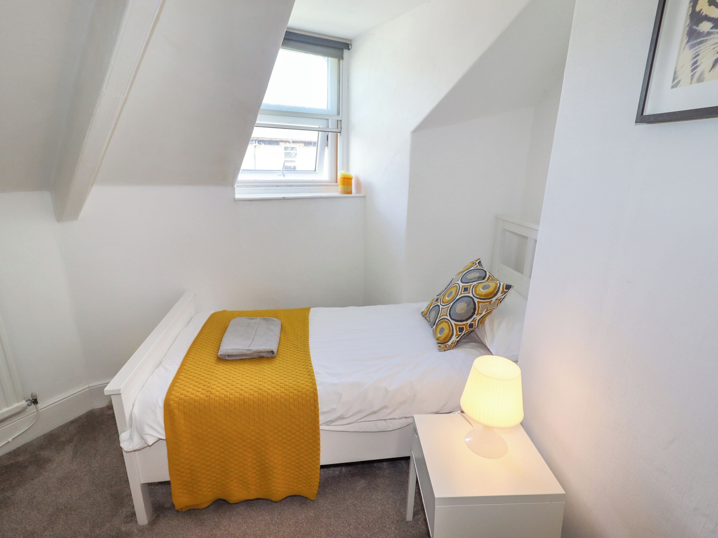 Une petite chambre à coucher, bien rangée, avec un lit simple, une lampe de chevet et une fenêtre.