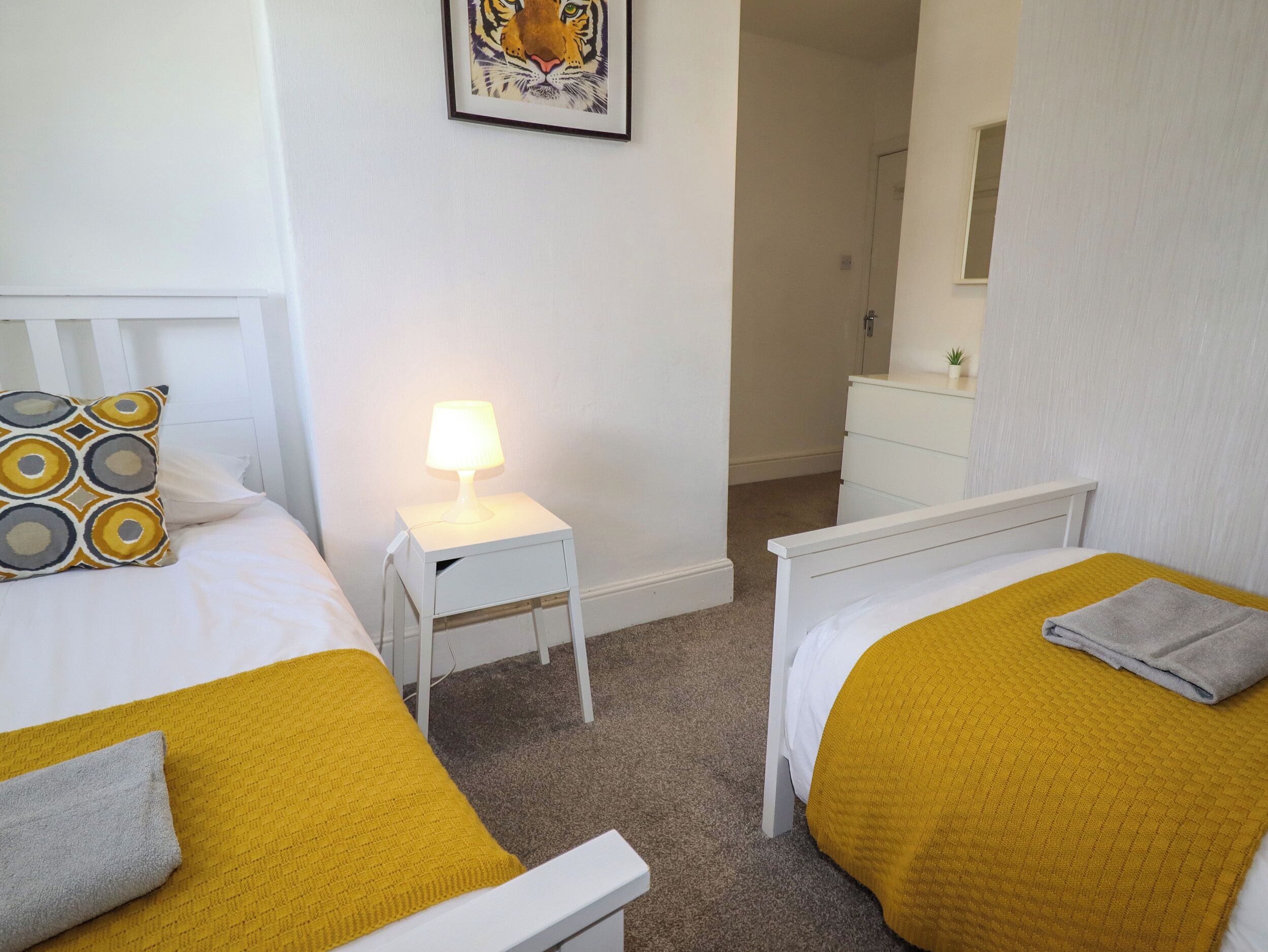 Et hotelværelse med to senge, et natbord med lampe, en lille kommode og et billede i ramme på væggen.