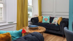 A living room with a dark sofa, a wooden coffee table, and a view of a body of water through a window with yellow curtains. - Sailors Rest (Dartmouth)