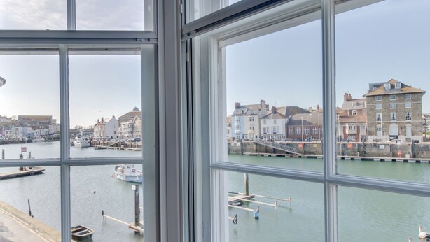 Blick durch ein Fenster auf einen Hafen mit vertäuten Booten, einer Pier und Gebäuden am Wasser.