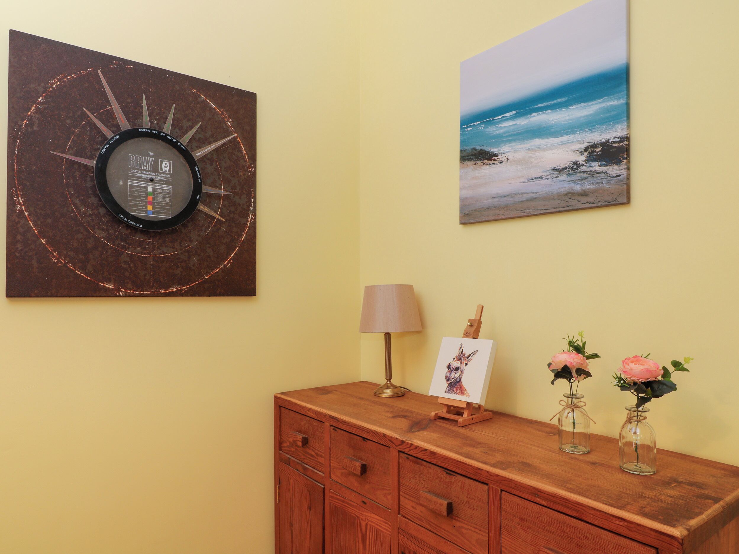 A wall with a rustic clock, a beach painting, a small easel with a picture, and a wooden cabinet with vases.