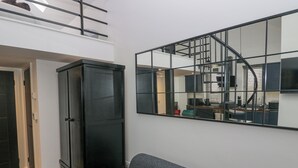 Una sala de estar moderna con un sofá gris, un mueble negro y un espejo grande que refleja la cocina y la escalera.