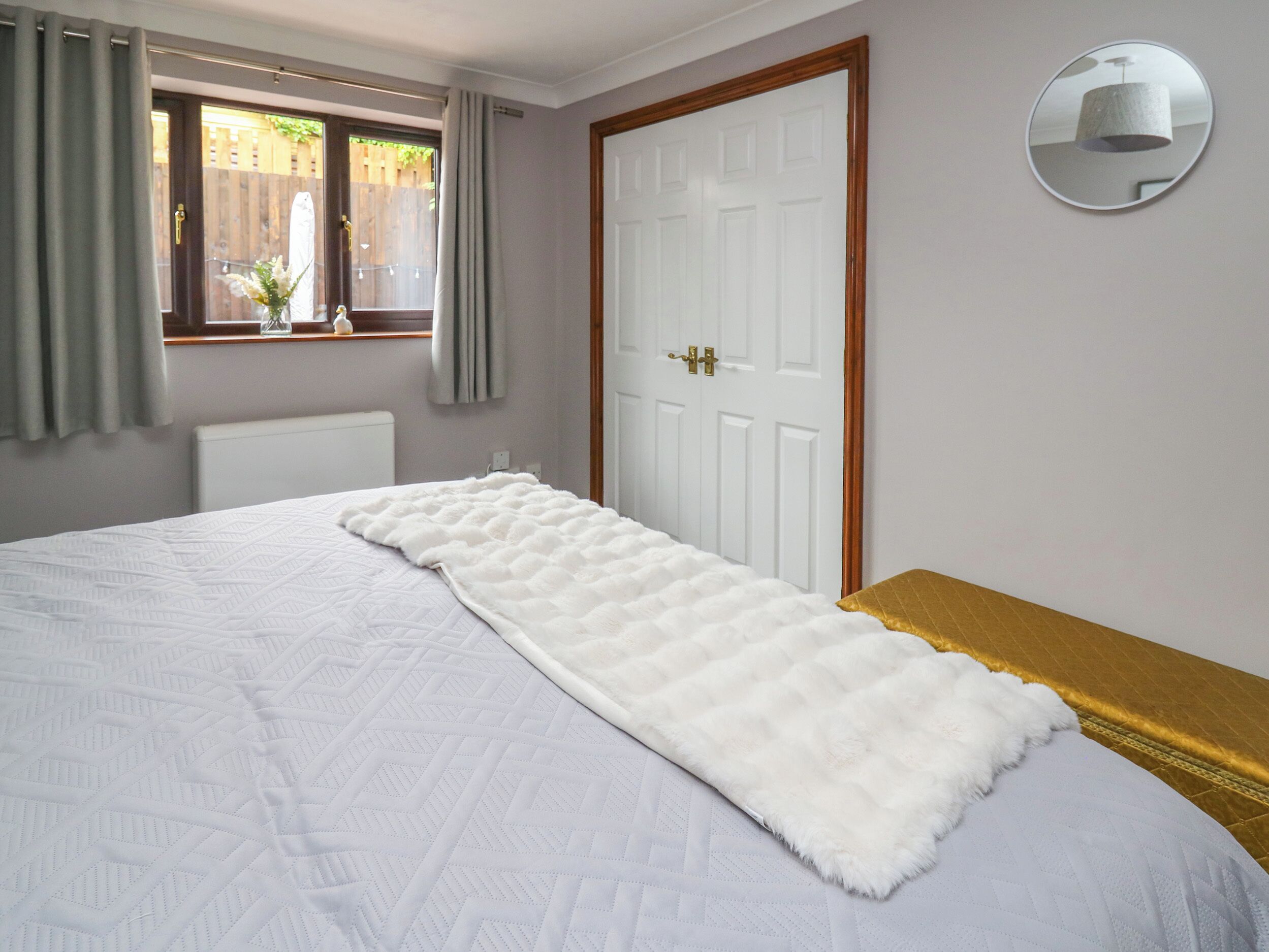 A bedroom with a bed, a window with curtains, a white door, and a round mirror on the wall.