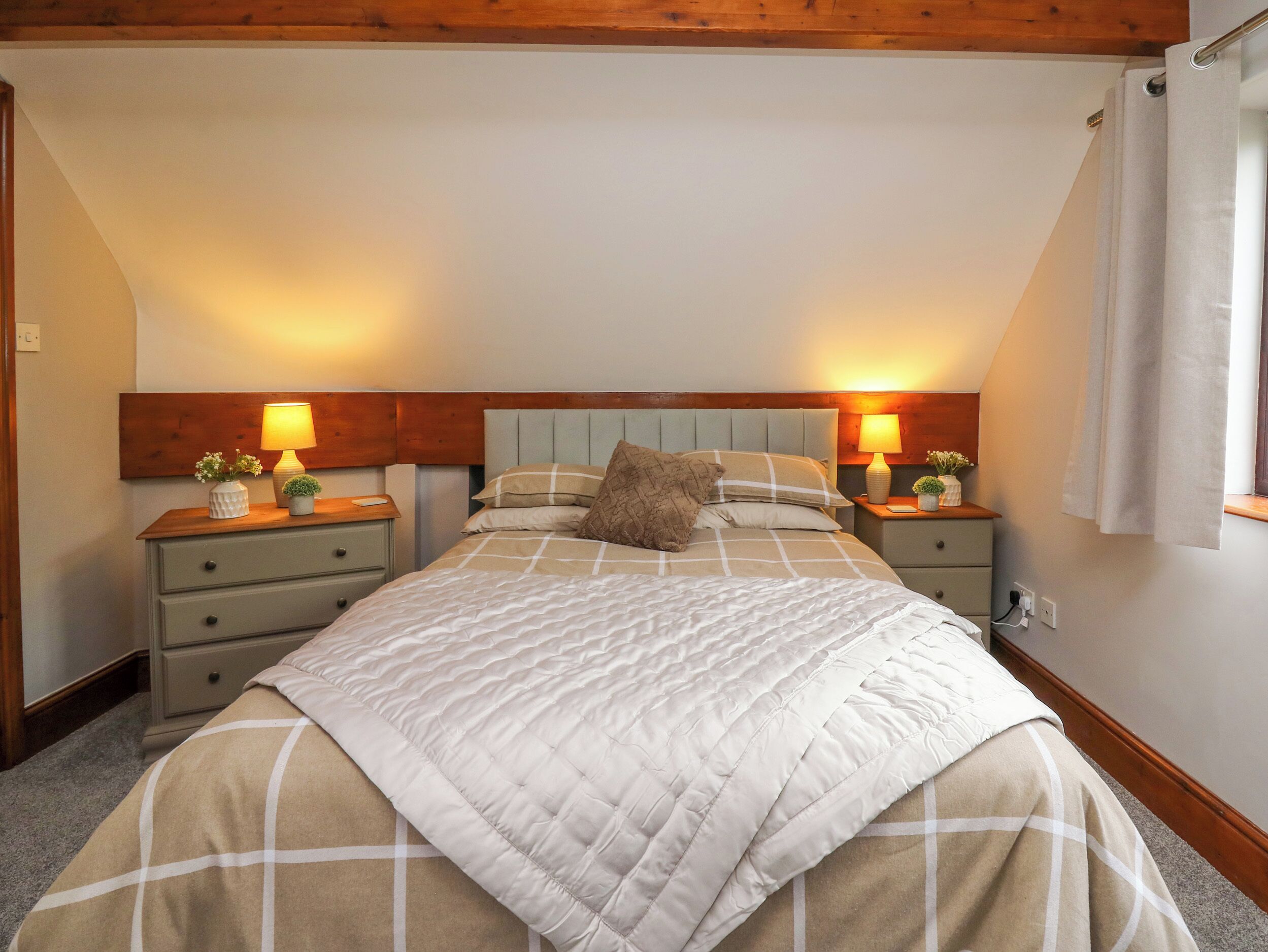 Ein Schlafzimmer mit einem großen Bett, zwei Nachttischen mit Lampen, einem hölzernen Kopfteil und einem Fenster mit Vorhängen.