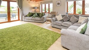 A spacious living room with two grey sofas, a green rug, wooden beams on the ceiling, and large windows.