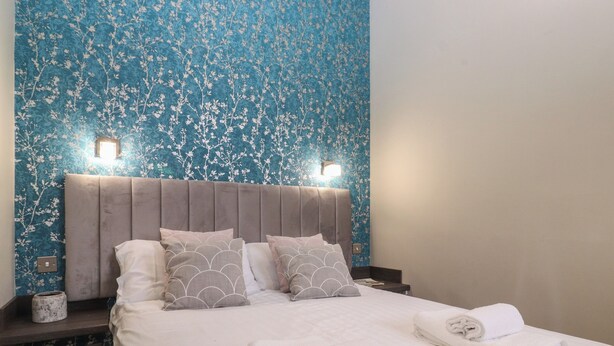Un lit bien fait, avec des draps blancs et deux serviettes enroulées, une tête de lit ornée d’un motif floral et une table de chevet avec une lampe et un petit récipient.
