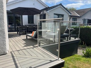 A glass-enclosed outdoor seating area with a table and chairs, a glass railing, and a view of a house in the background. - Yonder Lea (Windermere)