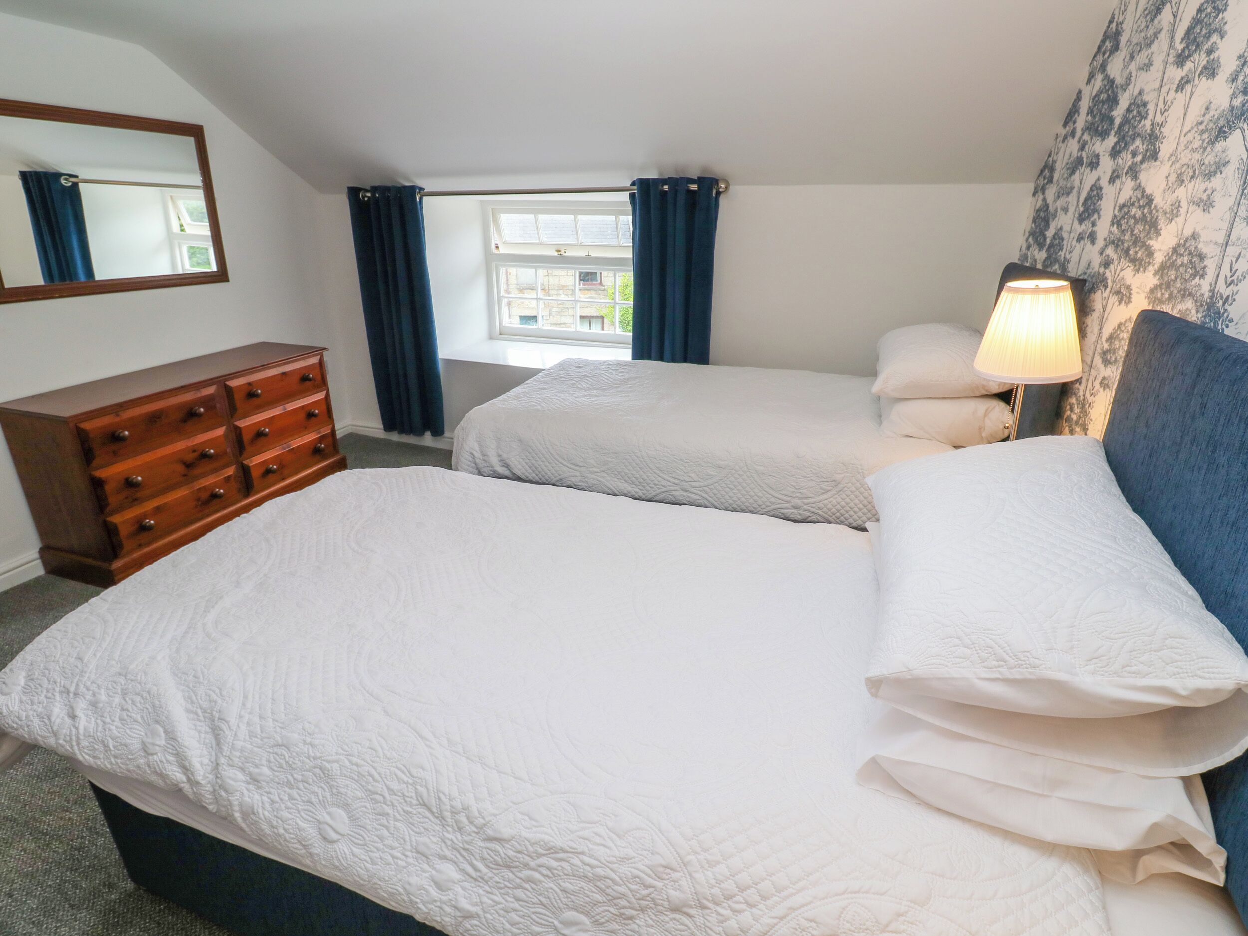 A bedroom with two beds, a wooden dresser, a window with curtains, and a lamp.