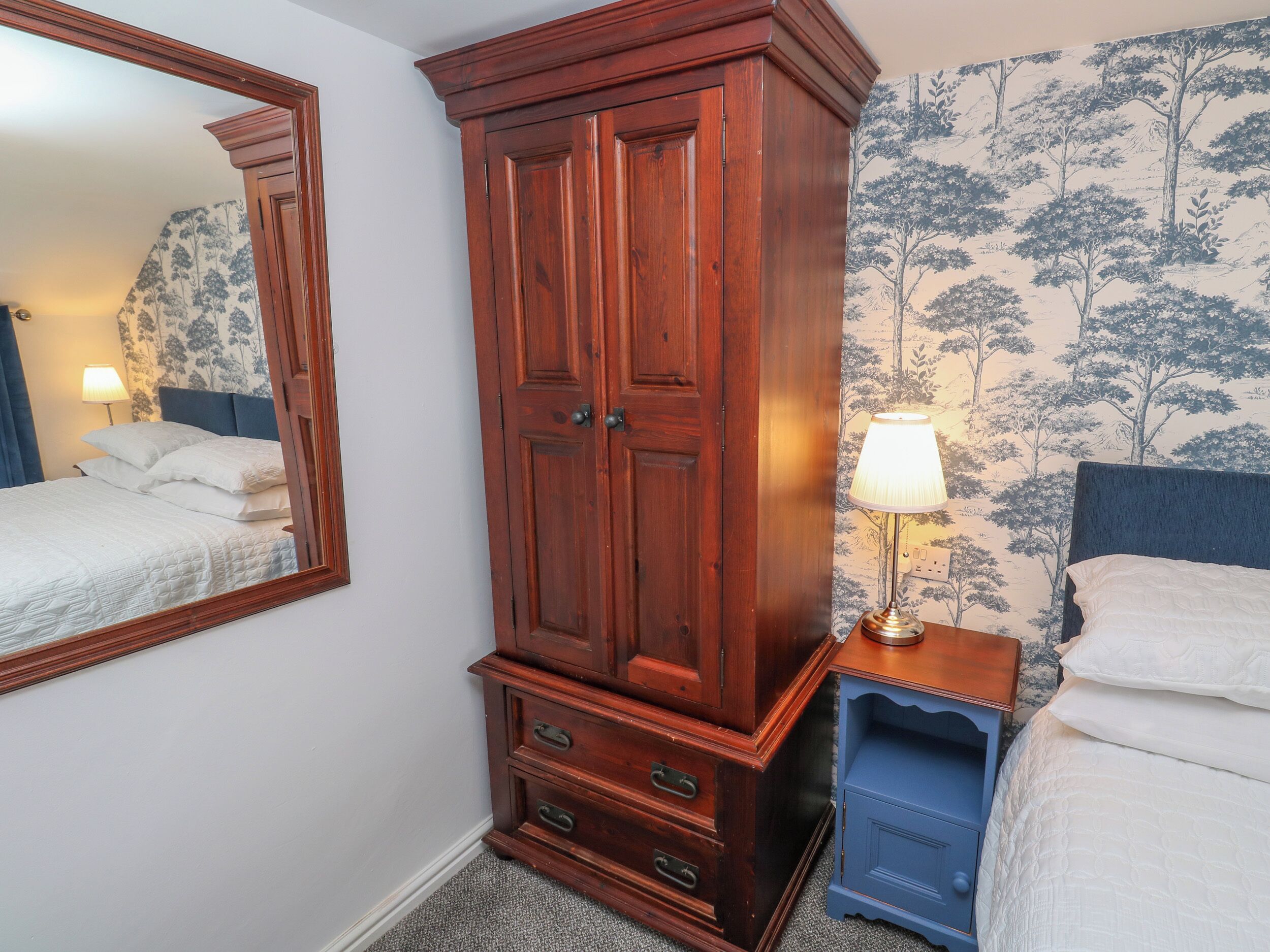 A bedroom with a large wooden wardrobe, a bed with white bedding, a bedside table with a lamp, and a mirror on the wall.