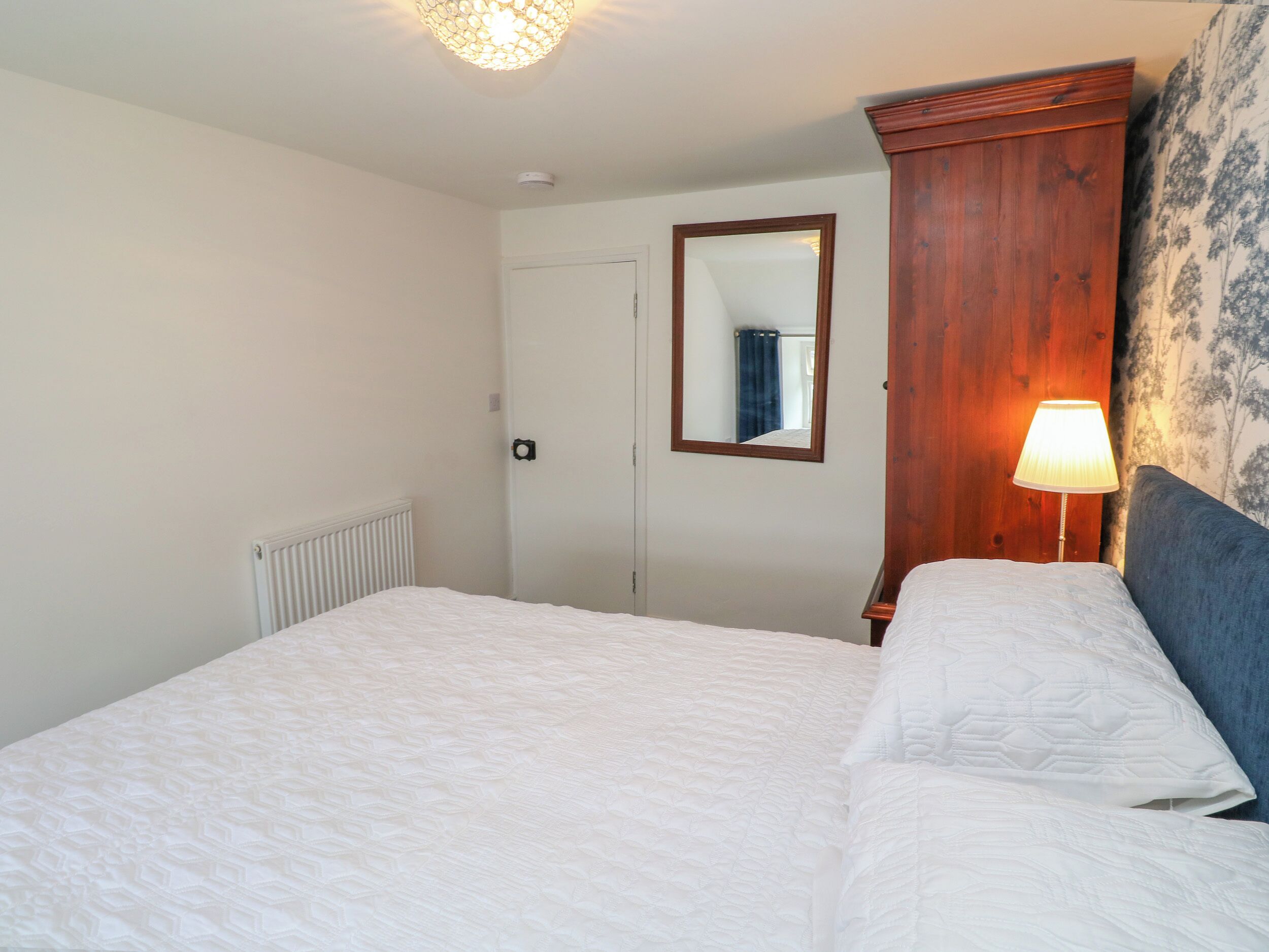 A bedroom with a bed, a wooden wardrobe, a mirror, a radiator, and a wall with a floral pattern.