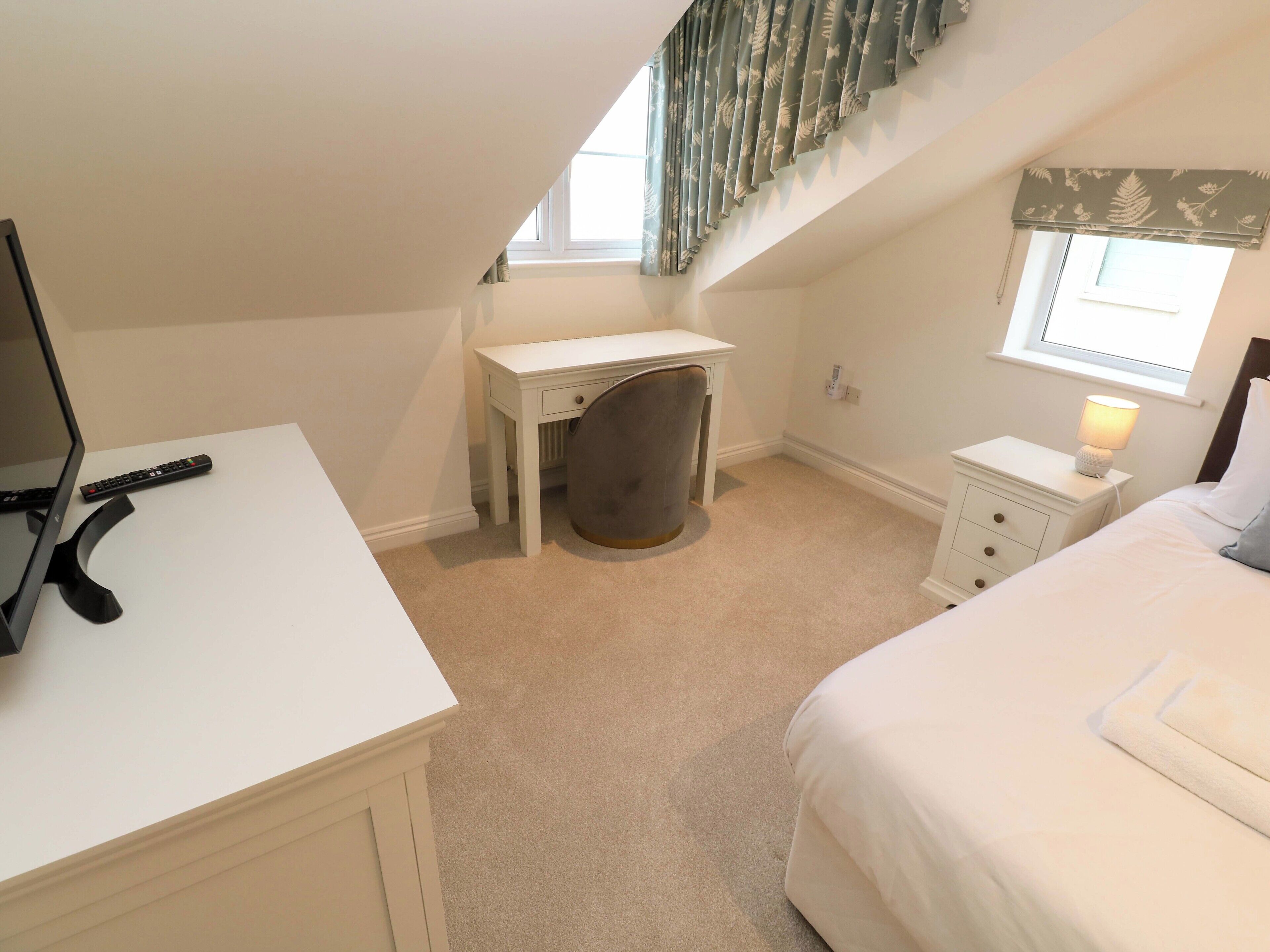 A bedroom with a white desk, a chair, a bed, a nightstand with a lamp, and a window with curtains.