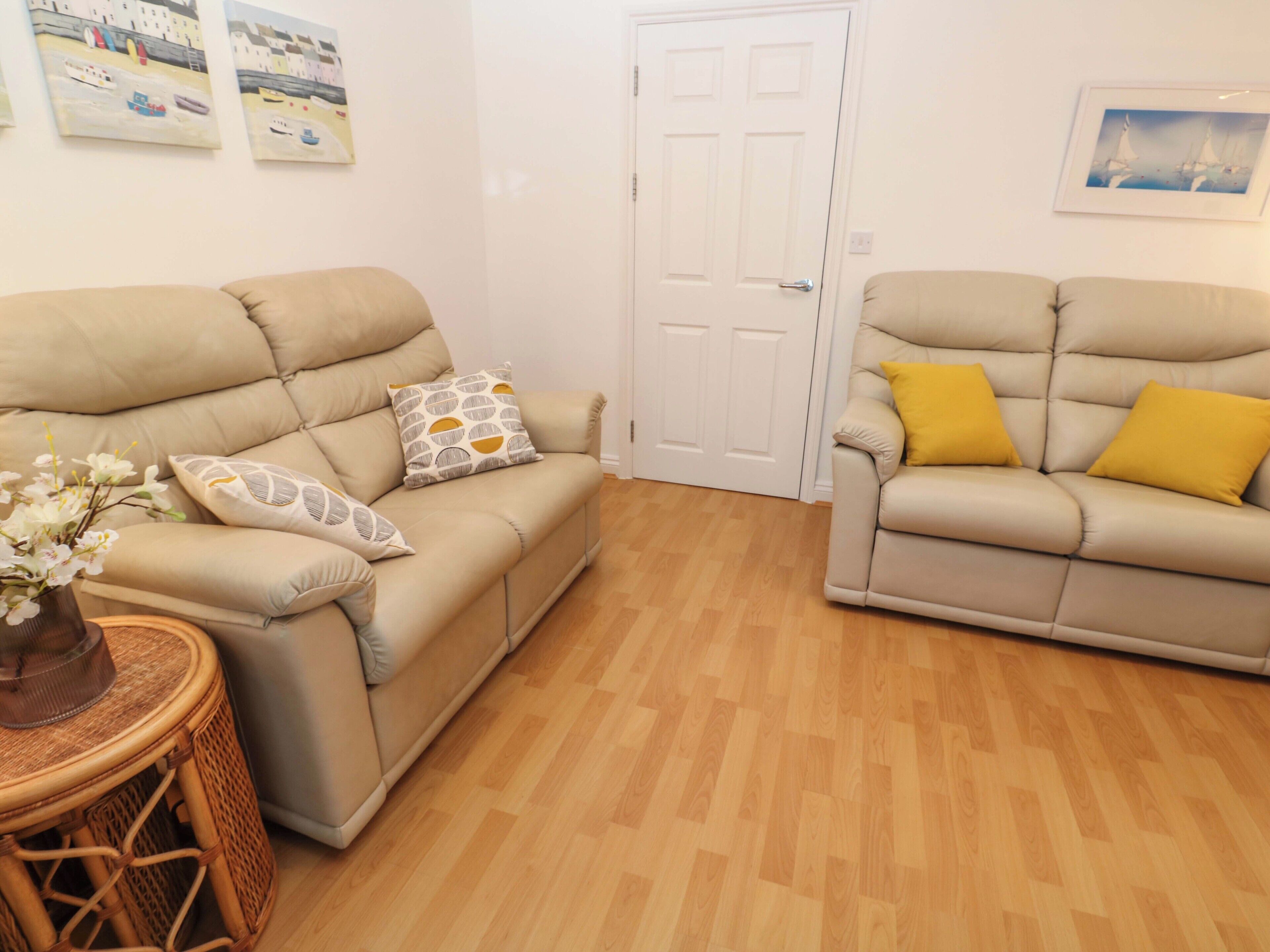 A living room with two beige sofas, a small round table with a vase of flowers, wooden flooring, and two doors.