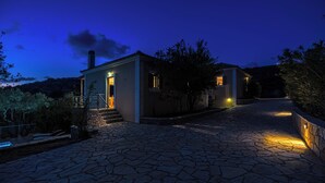 Uma trilha de pedras conduz a uma casa de dois andares com chaminé, cercada por árvores e um céu claro.