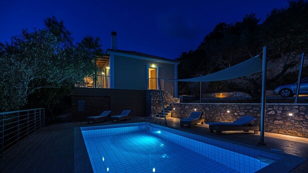 Piscina com espreguiçadeiras e parede de pedra, cercada por árvores e uma casa com entrada iluminada.