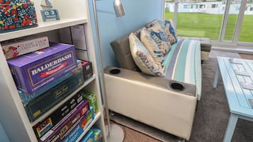 A room with a bookshelf filled with board games, a sofa with patterned cushions, a lamp, and a view of a grassy area through large windows.