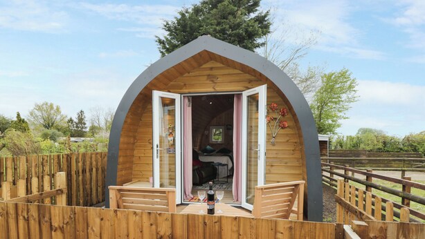 A wooden cabin with a curved roof, a wooden deck, and a glass door leading to a room with a bed and a table. - The Poddy (Swindon)
