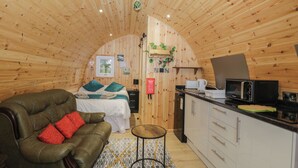 A cozy, wooden interior with a curved ceiling, a bed with teal bedding, a leather sofa with red cushions, a small round table, and a kitchenette with a microwave. - The Poddy (Swindon)